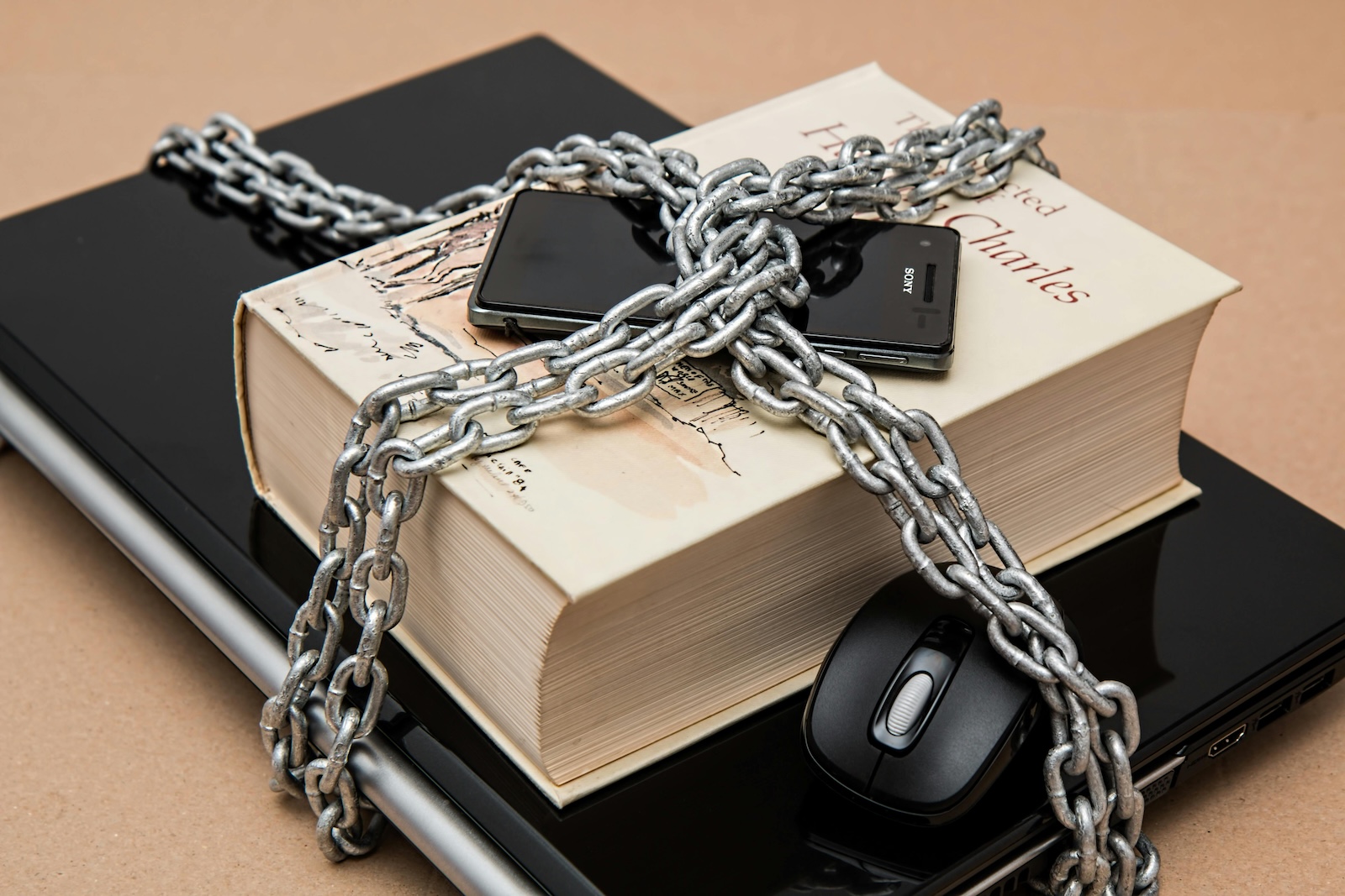 A laptop, book, mouse, and cellphone are wrapped up in a thick chain that is secured with a padlock.