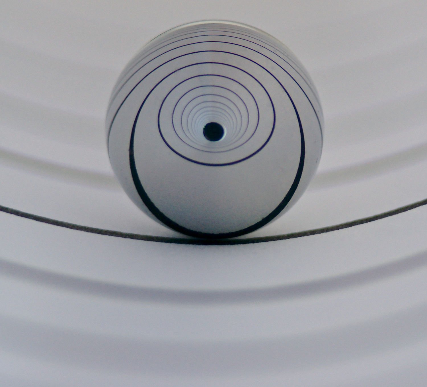 A small sphere is in the center of the frame with a curved line beneath it. Within the sphere, there are increasingly inward spirals culiminating in a dark hole in the center of the sphere.